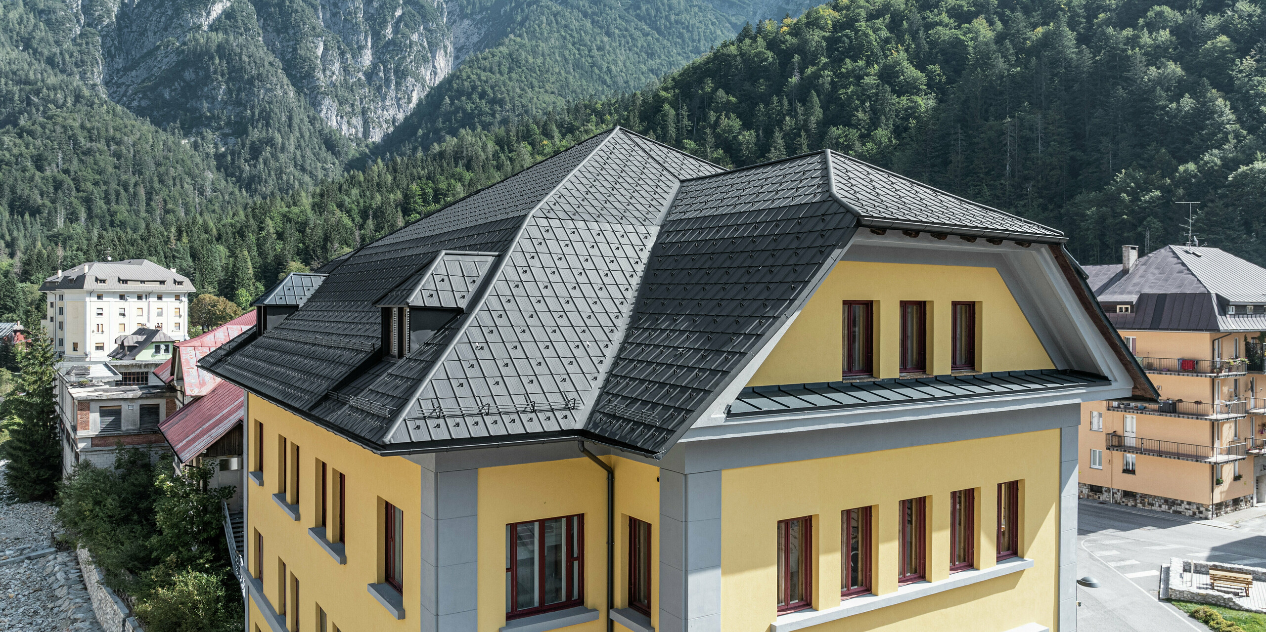 Bâtiment de musée historique Museo Cave del Predil rénové avec style avec des tuiles PREFA robustes en P.10 anthracite. La solution moderne en aluminium PREFA protège le bâtiment de manière fiable et durable contre les influences météorologiques alpines. En arrière-plan, l'impressionnant paysage de montagne avec ses pentes boisées souligne les exigences de robustesse et de résistance aux intempéries. Solution de toiture idéale de PREFA pour une utilisation dans les régions à fortes charges de neige et aux conditions climatiques exigeantes.