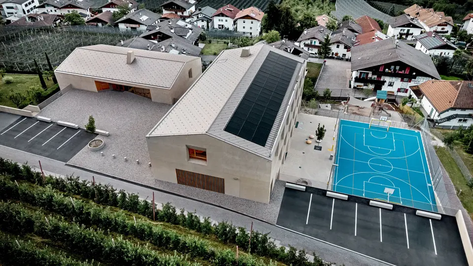 Vue aérienne du centre éducatif de Riffian, dans le Tyrol du Sud, comprenant un jardin d’enfants, une école primaire et un terrain de sport. Les losanges de toiture PREFA 44 × 44, dans le coloris P.10 Bronze, forment une toiture en aluminium précise et durable qui s’intègre harmonieusement au paysage alpin. Cette architecture moderne se distingue par sa résistance aux intempéries et par un design épuré, soigné jusque dans les moindres détails.