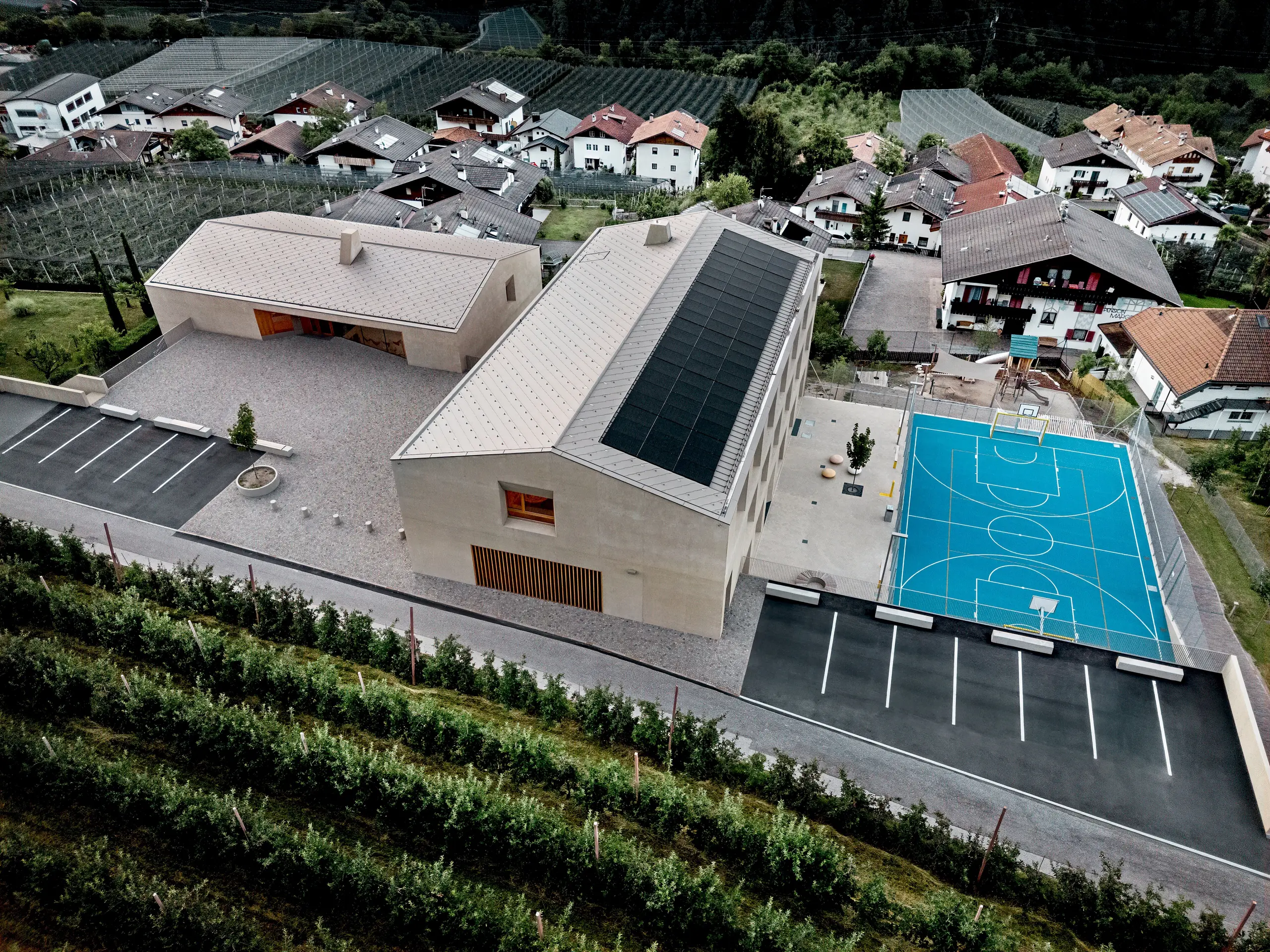 Vue aérienne du centre éducatif de Riffian, dans le Tyrol du Sud, comprenant un jardin d’enfants, une école primaire et un terrain de sport. Les losanges de toiture PREFA 44 × 44, dans le coloris P.10 Bronze, forment une toiture en aluminium précise et durable qui s’intègre harmonieusement au paysage alpin. Cette architecture moderne se distingue par sa résistance aux intempéries et par un design épuré, soigné jusque dans les moindres détails.