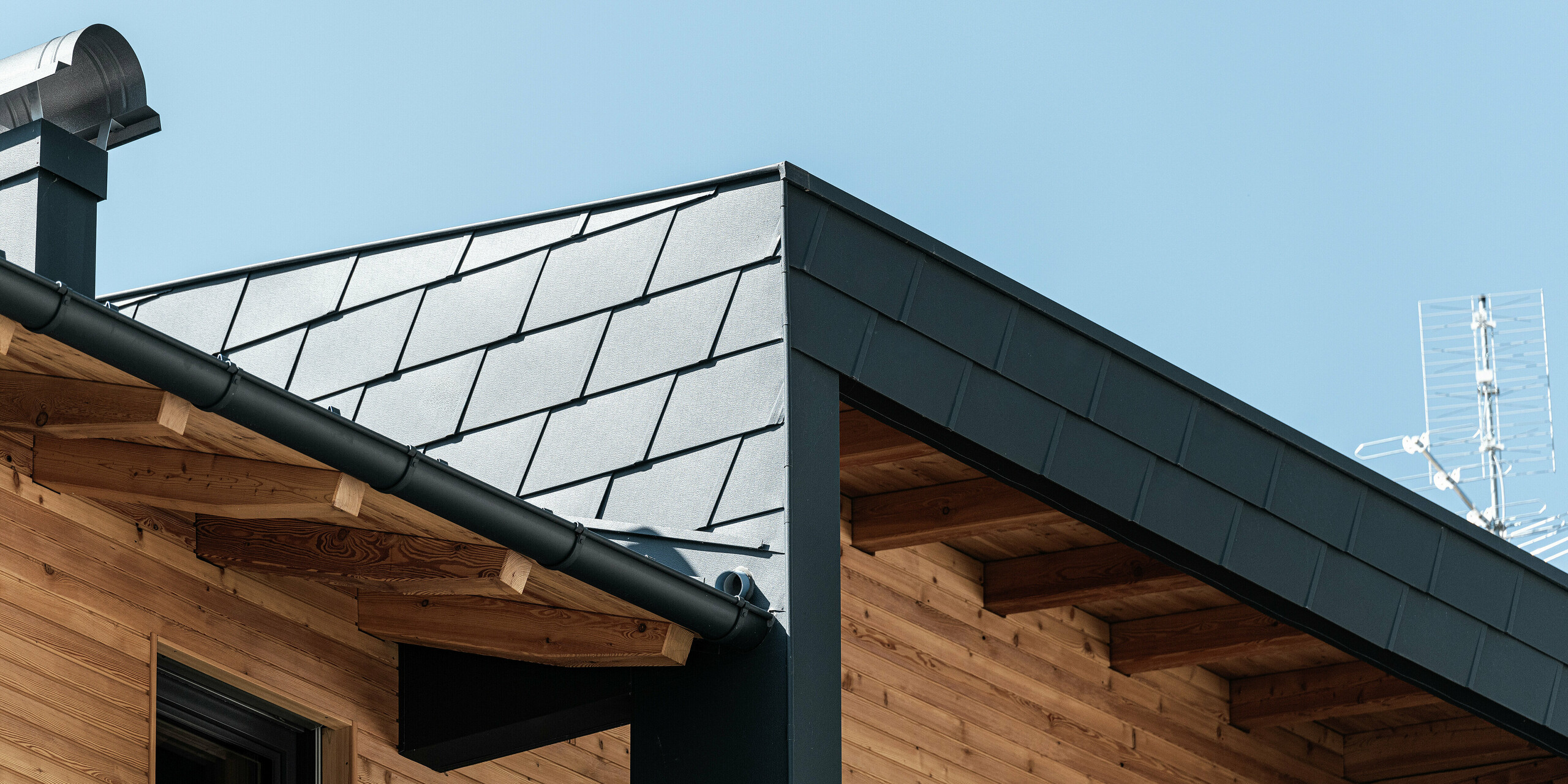 Vue détaillée de l'angle de toiture avec les DS.19 PREFA de couleur P.10 anthracite et du revêtement de façade du balcon couvert. La combinaison de l'aluminium et du bois apporte des accents esthétiques.