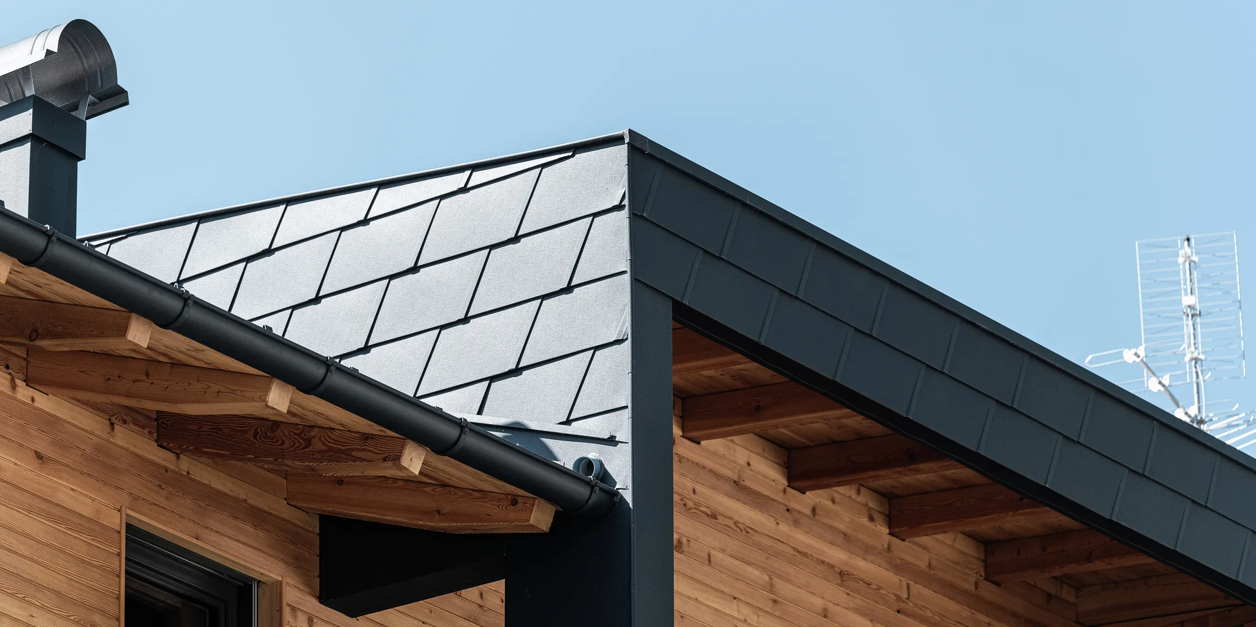 Vue détaillée de l'angle de toiture avec les DS.19 PREFA de couleur P.10 anthracite et du revêtement de façade du balcon couvert. La combinaison de l'aluminium et du bois apporte des accents esthétiques.