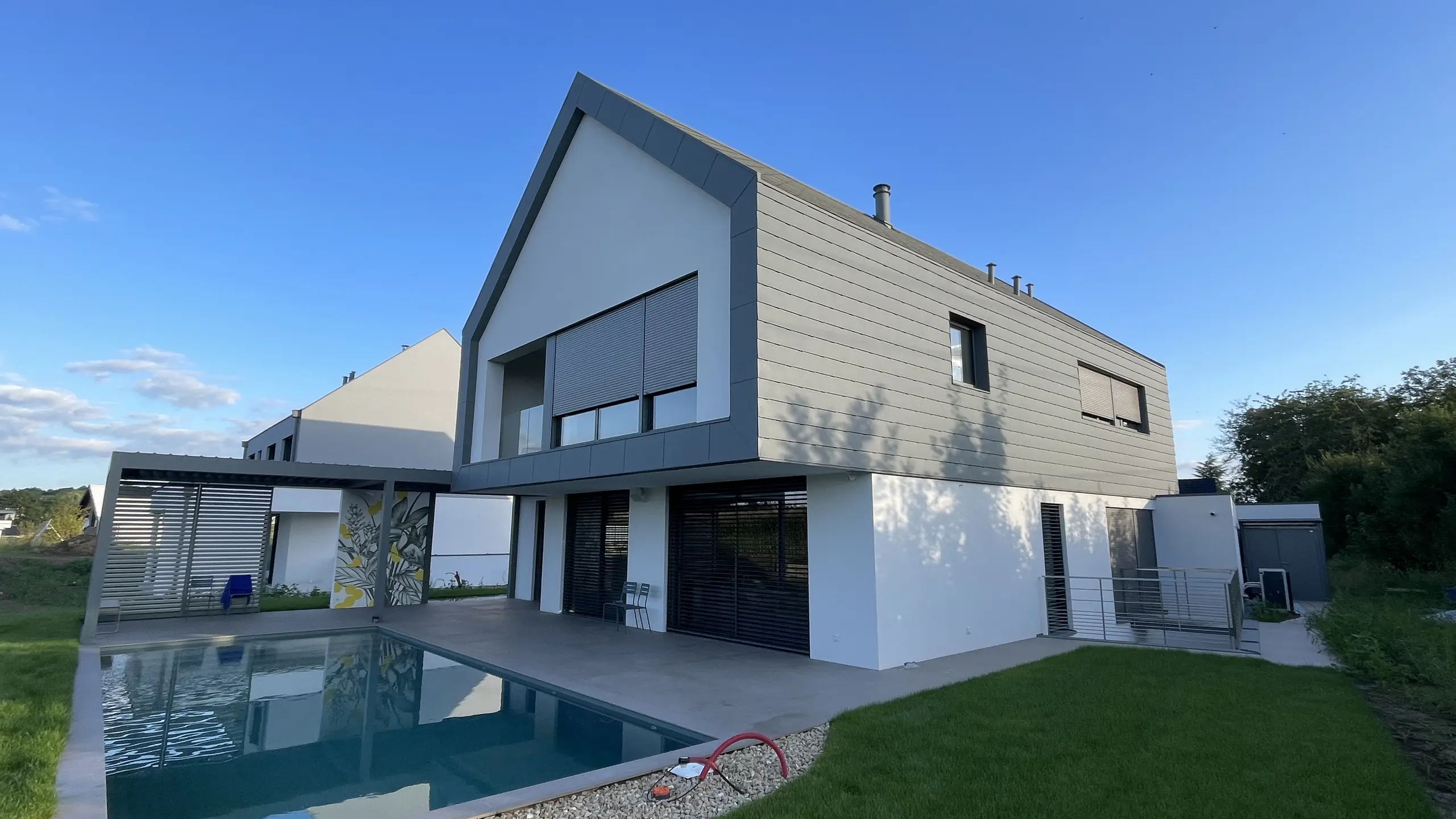 Vue d'ensemble de cette nouvelle maison individuelle dont la toiture et la façade contemporaine ont été recouvertes de bardeaux en aluminium PREFA. On aperçoit une jolie piscine en premier plan.