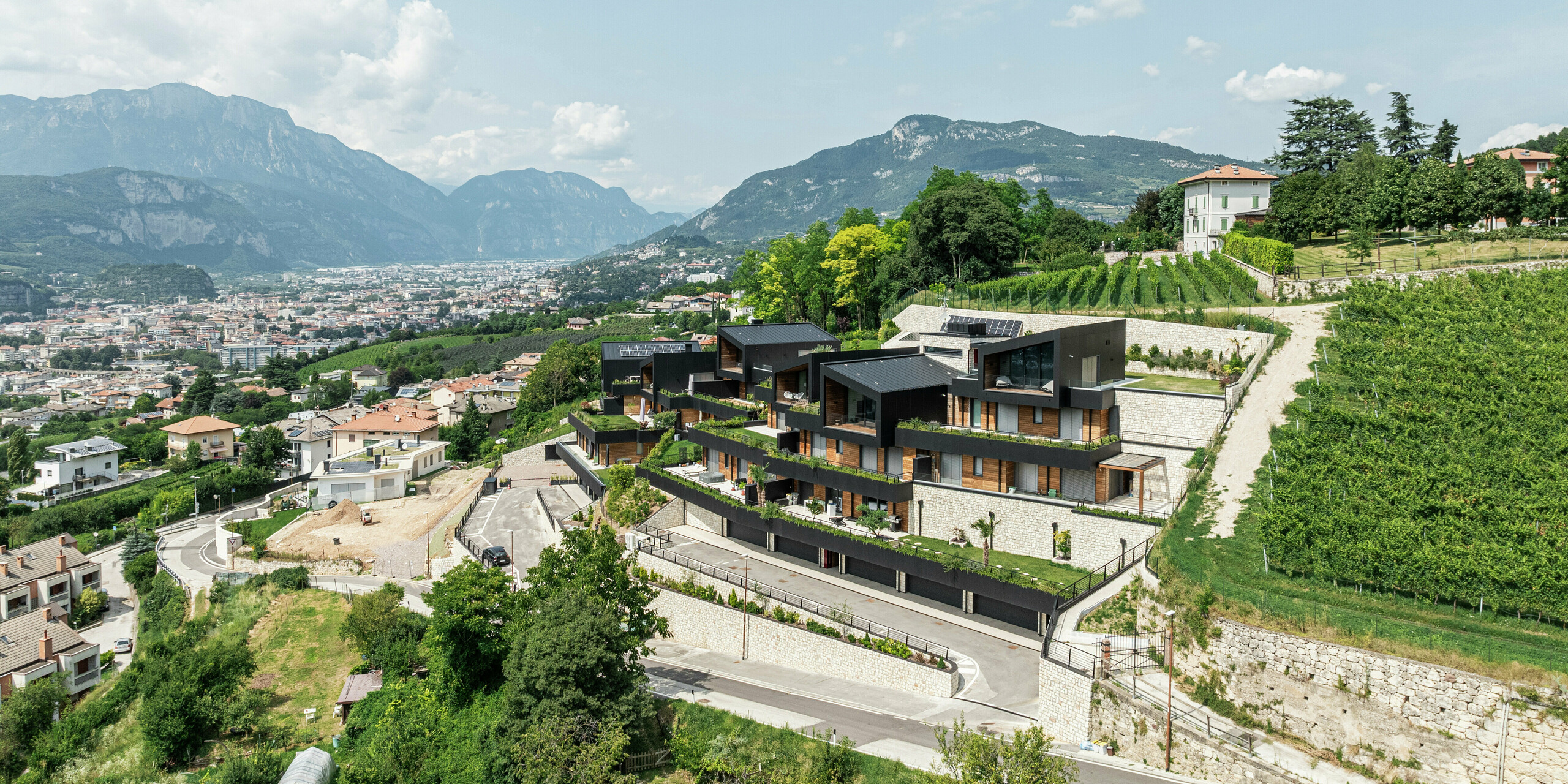 Emplacement spectaculaire à flanc de colline avec vue sur Trente : le complexe résidentiel exclusif s'intègre parfaitement dans les vignobles environnants et le paysage vallonné. L'architecture moderne combine la pierre naturelle, le bois et la façade en aluminium PREFA durable en P.10 Noir pour créer une unité élégante et résistante aux intempéries. Le toit PREFALZ en P.10 Noir assure une protection et une durabilité maximales. Des terrasses spacieuses et des espaces verts soulignent le concept de vie durable et créent une interaction harmonieuse entre la nature et la construction moderne.