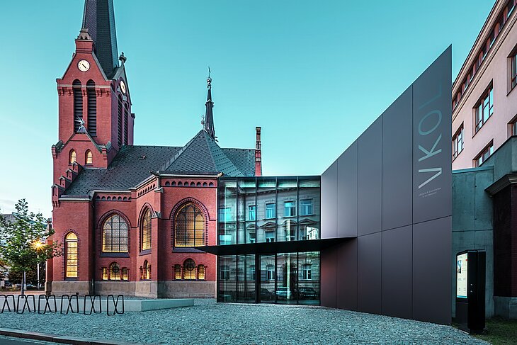 L'image révèle l'interaction remarquable entre l'église rouge historique et le nouveau bâtiment moderne d'Olomouc. L'église, avec ses fenêtres gothiques caractéristiques et son clocher imposant, constitue la colonne vertébrale historique de l'ensemble, tandis que l'extension moderne, avec son élégante façade en panneaux composites en aluminium PREFABOND noirs, représente une réponse architecturale contemporaine. De larges baies vitrées offrent des vues sur l'intérieur du nouveau bâtiment, où les reflets dans les fenêtres créent un jeu subtil entre ancien et nouveau. La précision de la fabrication des panneaux PREFABOND souligne l'esthétique moderne, tandis que l'intégration harmonieuse dans le contexte historique impressionne autant par sa fonctionnalité que par son esthétique.