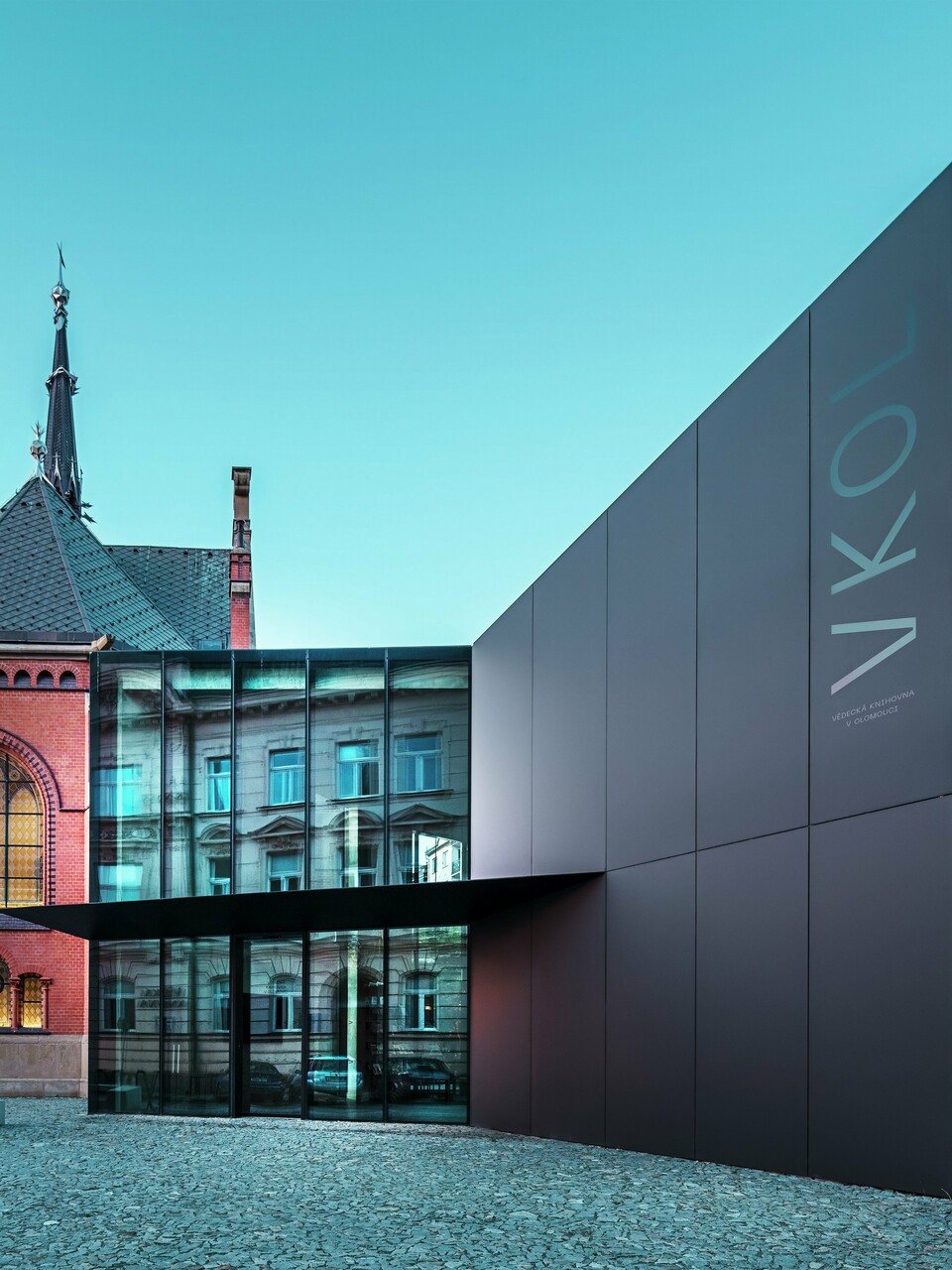 L'image révèle l'interaction remarquable entre l'église rouge historique et le nouveau bâtiment moderne d'Olomouc. L'église, avec ses fenêtres gothiques caractéristiques et son clocher imposant, constitue la colonne vertébrale historique de l'ensemble, tandis que l'extension moderne, avec son élégante façade en panneaux composites en aluminium PREFABOND noirs, représente une réponse architecturale contemporaine. De larges baies vitrées offrent des vues sur l'intérieur du nouveau bâtiment, où les reflets dans les fenêtres créent un jeu subtil entre ancien et nouveau. La précision de la fabrication des panneaux PREFABOND souligne l'esthétique moderne, tandis que l'intégration harmonieuse dans le contexte historique impressionne autant par sa fonctionnalité que par son esthétique.