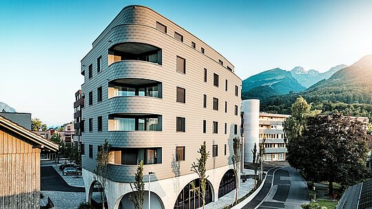 Vue oblique de la Bernerhaus à Buchs, reconnaissable à sa forme triangulaire caractéristique et à ses angles légèrement arrondis. La façade continue en bardage PREFA gris souris P.10 enveloppe le bâtiment comme une peau d'aluminium à écailles et souligne les lignes fluides des balcons. Au-dessus du socle en béton massif orné d'élégantes arcades, la façade en aluminium PREFA, légère et résistante aux intempéries, affirme une présence remarquable. Posée avec précision, durable et nécessitant peu d'entretien, elle s'intègre harmonieusement au paysage alpin et constitue une véritable déclaration architecturale.