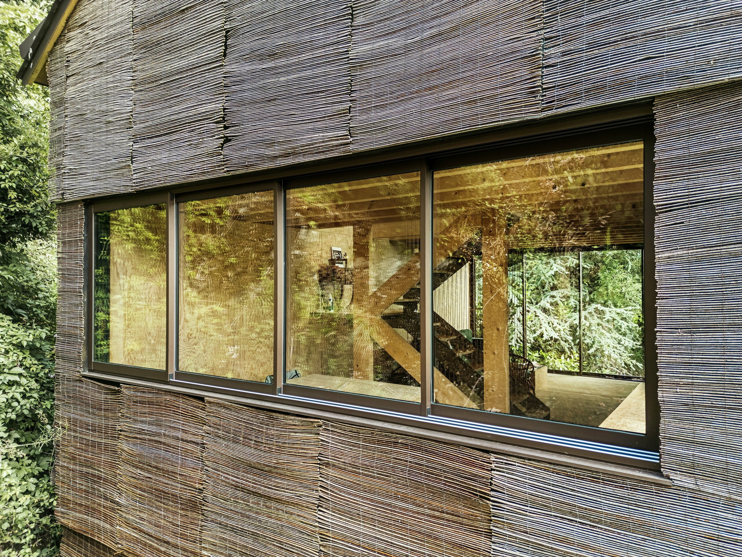 Détail d'une fenêtre avec un cadre en aluminium moderne et une façade en bois de saule sur une maison en France – conception durable avec des bardeaux de toiture PREFA et l'environnement naturel.