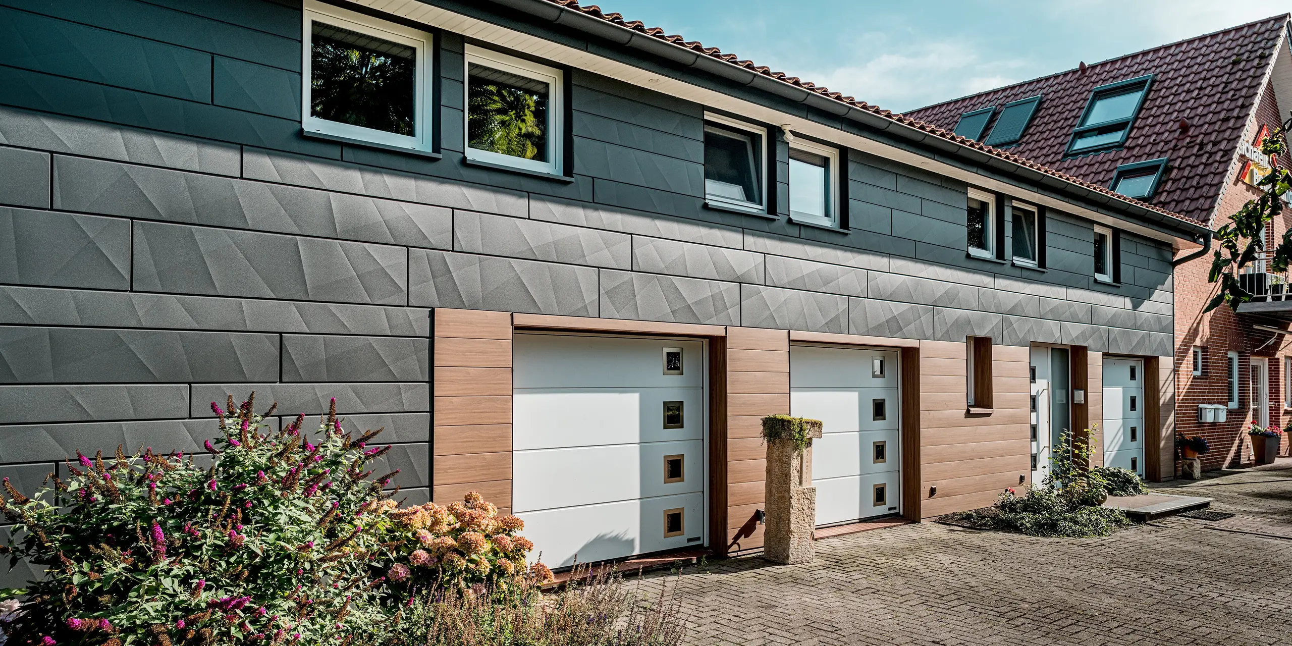 Vue en perspective d'une maison individuelle rénovée, dotée d'une façade moderne en aluminium PREFA et d'une structure horizontale épurée. Les panneaux Siding.X PREFA grand format couleur P.10 gris sombre, avec leurs arêtes longitudinales et transversales marquées, attirent particulièrement le regard et créent un jeu d'ombres et de lumières saisissant. Dans la partie inférieure de la façade, les panneaux Siding PREFA, couleur noyer foncé, à l'élégant aspect bois, apportent une touche chaleureuse et accueillante autour des portes de garage et des entrées. Les portes et les encadrements de fenêtres blancs créent des contrastes marqués. Durables, faciles d'entretien et résistants aux intempéries, les panneaux en aluminium PREFA font de cette rénovation de façade un exemple remarquable d'architecture moderne et durable.