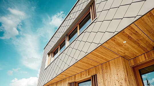 Le gros plan de la crèche d'Alzenau met en valeur la précision d'exécution et la qualité de l'association des losanges de façade 44x44 PREFA couleur bronze et du bois naturel. La structure diagonale de la façade en aluminium crée un motif d'écailles saisissant qui s'harmonise avec les persiennes verticales en bois entourant les fenêtres et le soffite en bois finement travaillé. La prise de vue en contre-plongée souligne la précision des lignes et le savoir-faire du bardage ventilé. Cette vue détaillée met en évidence la robustesse et la durabilité des produits PREFA, idéaux pour les projets architecturaux durables qui allient esthétique, fonctionnalité et responsabilité écologique au plus haut niveau.