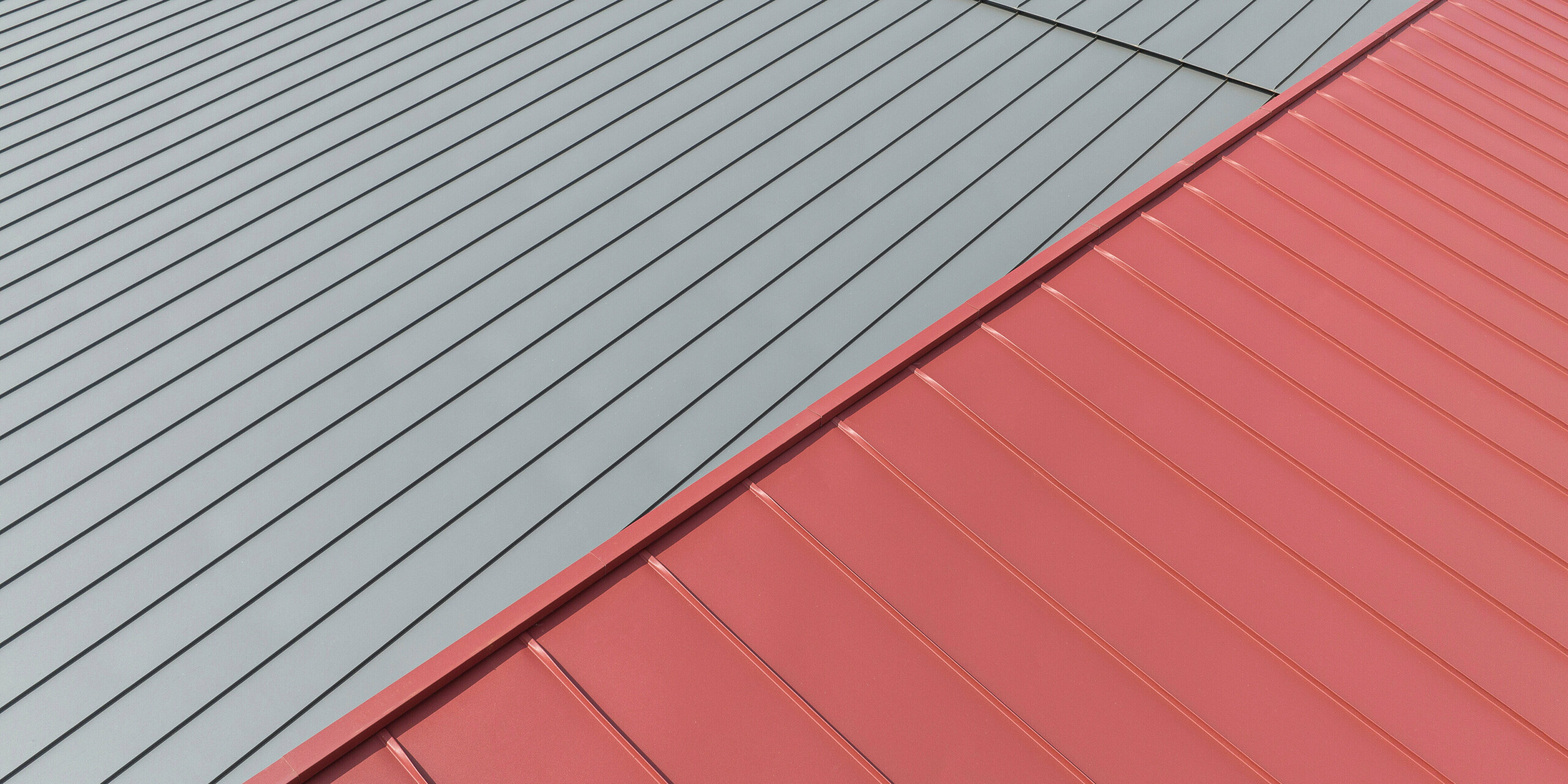 La photo montre deux sections d'un toit recouvert de PREFALZ. La plus grande partie est d'un gris sombre froid et la plus petite partie est d'un rouge oxyde vif. Les deux sections de toit ont un profil distinct commun aux toitures à joints debout en aluminium, ce qui indique une construction robuste et une conception durable. Le contraste entre les deux tons représente un design moderne et esthétique et suggère que la couleur est utilisée pour identifier différentes zones.