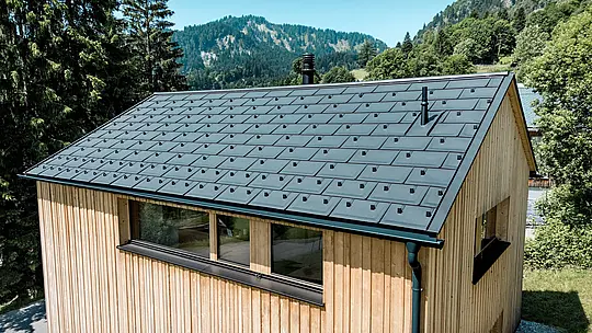 Sous la lumière de l’été alpin, le toit en aluminium noir PREFA de la maison Immi s’impose avec élégance. Les tuiles solaires intégrées à fleur de toiture forment, avec les R.16 en P.10 noir, une couverture homogène aux lignes épurées. Léger et robuste, le système en aluminium produit de l’énergie solaire et protège efficacement la maison en bois contre les conditions météorologiques exigeantes du milieu alpin ainsi que les fortes charges de neige.