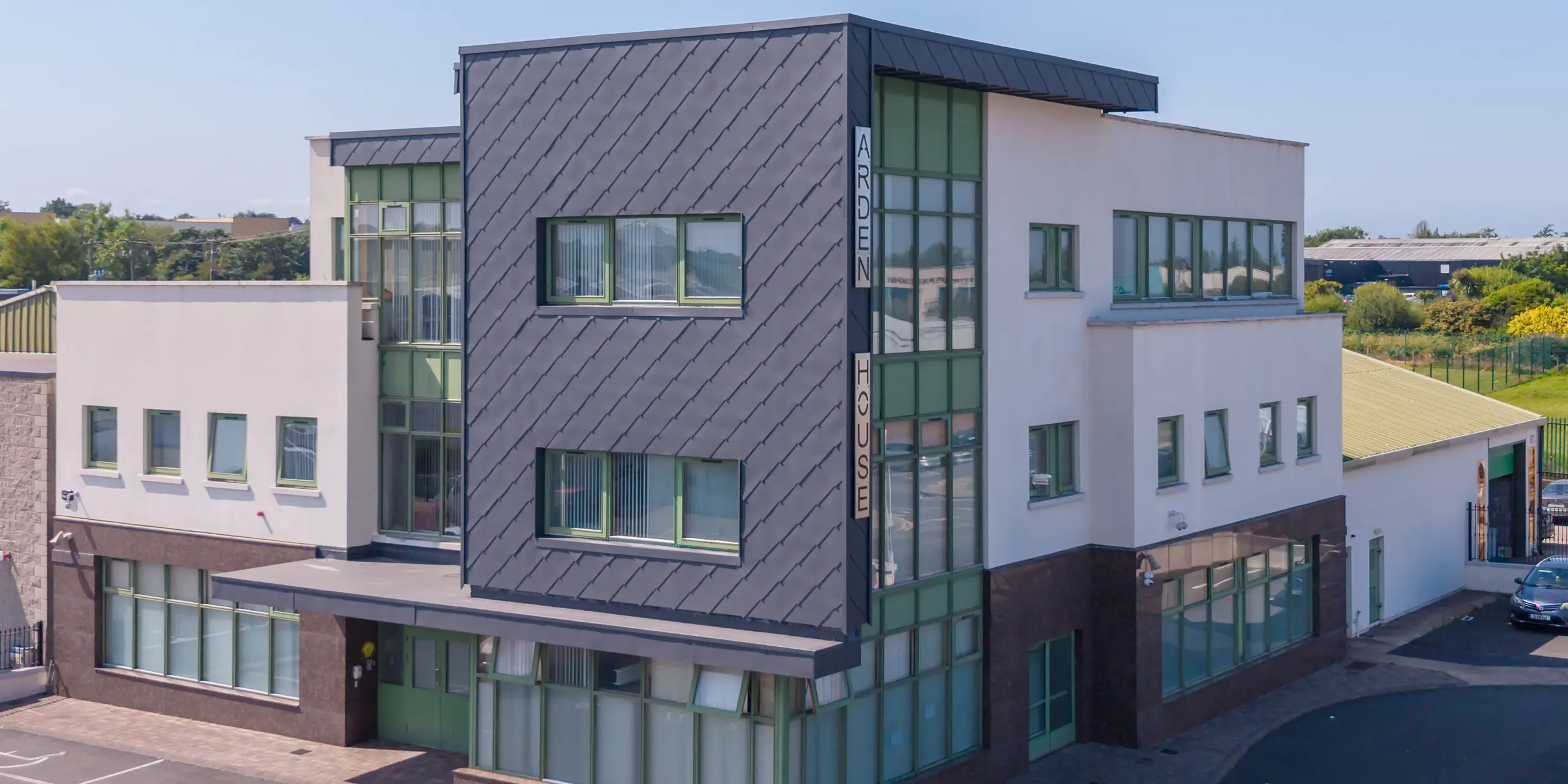 Markanter Gebäudekubus des Arden House, Zentrum für psychische Gesundheit von Kindern und Jugendlichen, in Wexford mit PREFA Wandraute 44x44 in P.10 Dunkelgrau. Die dynamische Rautenstruktur betont die klare Architektur, während die grünen Fenster Akzente setzen. Die langlebige und korrosionsbeständige Aluminiumfassade überzeugt durch Wartungsarmut und hohe Widerstandsfähigkeit.