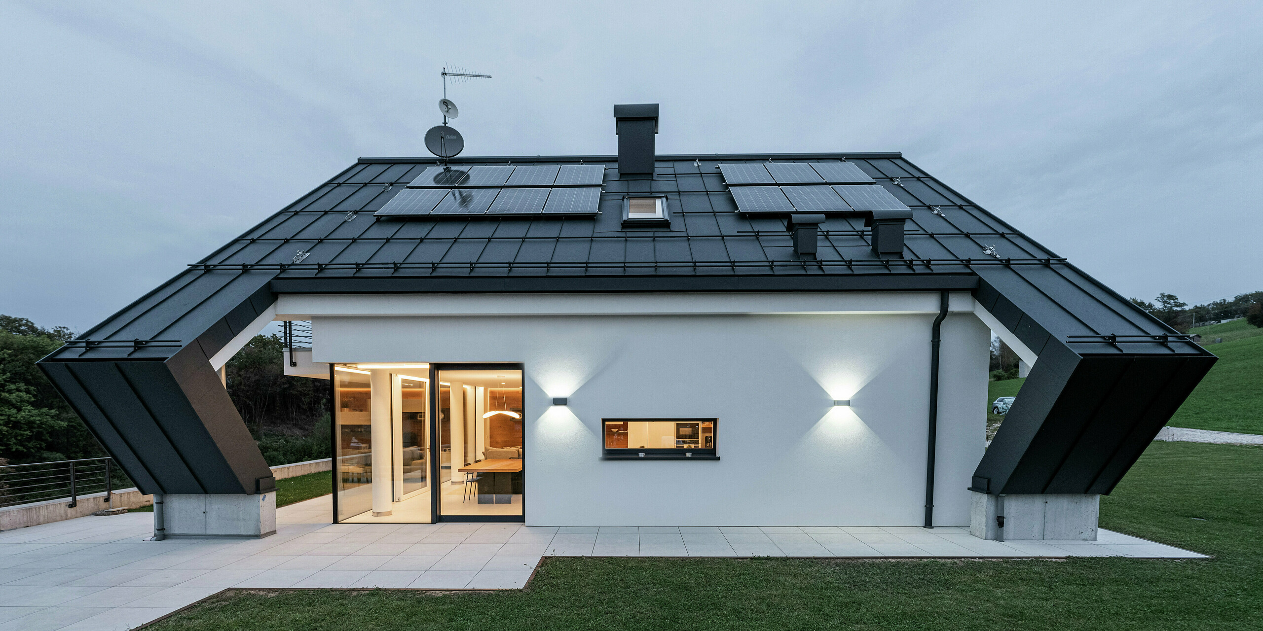 Maison familiale moderne à Borgo Valbelluna, en Italie, avec un toit à pignon remarquable en PREFALZ en P.10 anthracite. La couverture de toit en aluminium durable offre une protection optimale contre les éléments et impressionne par ses options d'installation flexibles. Les modules photovoltaïques intégrés utilisent efficacement la surface du toit pour générer de l’énergie durable. La combinaison du toit en aluminium de couleur anthracite et de la façade blanche ainsi que le porte-à-faux frappant du toit à pignon soulignent le design élégant et intemporel du bâtiment.