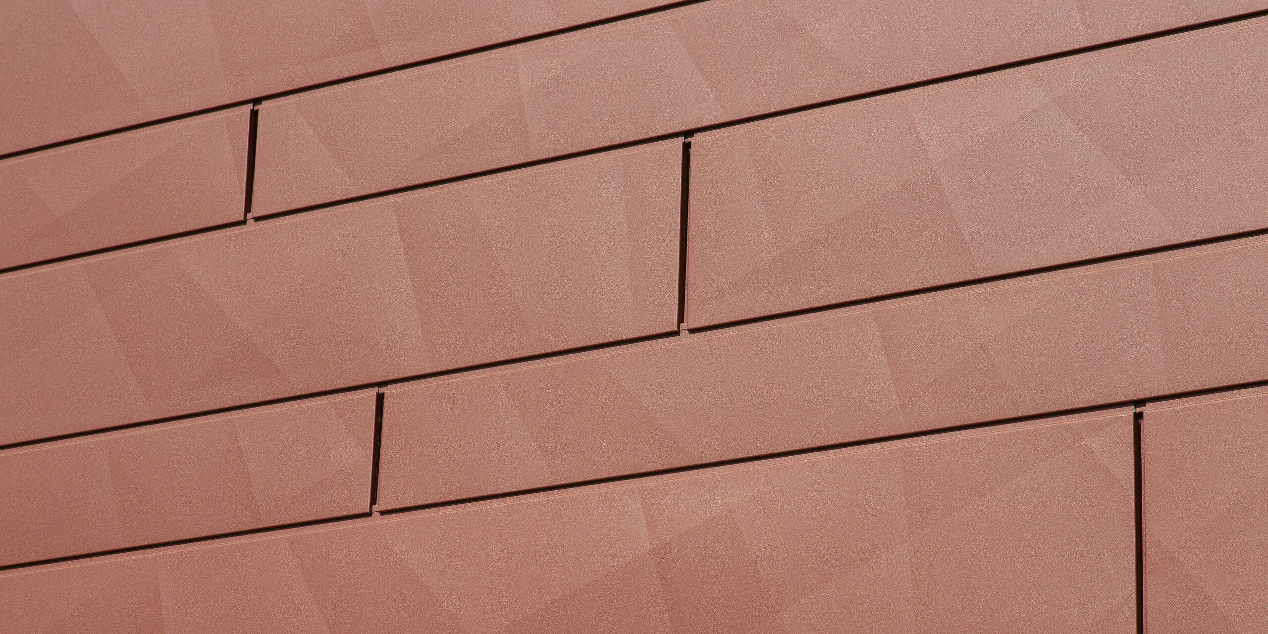 Gros plan sur les panneaux de façade Siding.X PREFA en rouge oxyde : le bord distinctif crée une structure géométrique saisissante qui reflète la lumière différemment selon l’heure de la journée. Le revêtement en poudre P.10 de haute qualité garantit une brillance durable des couleurs et une résistance maximale – idéal pour des rénovations de façade durables et sans entretien.