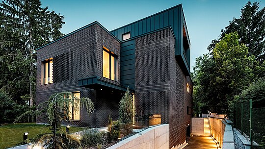La photographie révèle, au crépuscule, l'extérieur moderne et structuré d'une villa multigénérationnelle à Budapest. Les façades en briques brun-rouge foncé contrastent avec le bardage en aluminium PREFALZ P.10 anthracite, aux lignes verticales précises, qui souligne l'architecture du bâtiment. La lumière chaleureuse des fenêtres lui confère profondeur et atmosphère. L'allée menant au garage souterrain, subtilement éclairée, longe élégamment la façade et met en valeur la qualité, la durabilité et la résistance aux intempéries de l'alliance de la brique et de l'aluminium PREFA.
