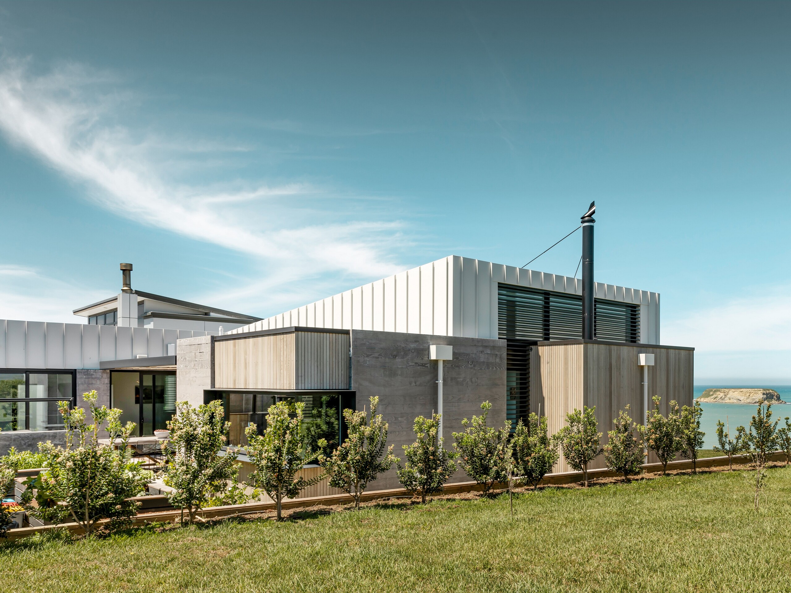 Seitlicher Blick auf das Whitecaps House und dessen Materialkombination aus Aluminium, Holz und Beton. Davor eine Begrünung, im Hintergrund sind Motunau Island und der Pazifik sichtbar.