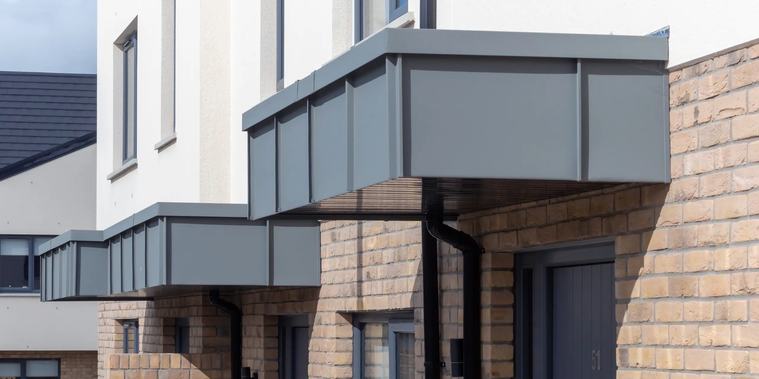 L'image présente les auvents d'entrée remarquables des maisons mitoyennes de Churchfields, entièrement revêtus de PREFALZ couleur P.10 gris souris. Les lignes épurées et droites des surfaces en aluminium créent un contraste moderne avec la chaleur de la brique et confèrent à l'ensemble une apparence uniforme et haut de gamme. La surface robuste de PREFA est résistante aux intempéries, inaltérable et ne nécessite pratiquement aucun entretien – idéale pour les bâtiments résidentiels durables. Le gros plan met en valeur la précision de la pose et l'intégration harmonieuse du revêtement en aluminium à l'architecture.