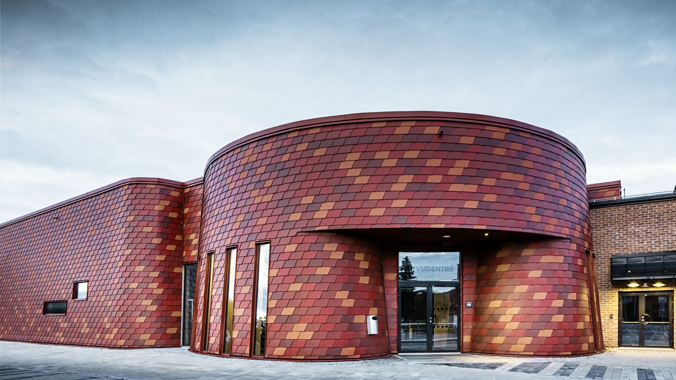 Patinoire à toit plat et façade incurvée ; la façade est revêtue de bardeaux de façade en aluminium de PREFA dans différentes nuances de rouge - rouge oxyde, rouge tuile, brun rouge, rouge brun