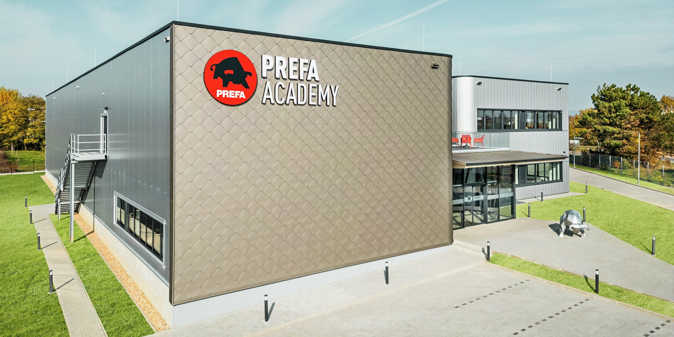 La PREFA Academy de Törökbálint se présente avec une façade saisissante composée de losanges de façade PREFA 44 × 44 en bronze et de Sidings PREFA en argent métallisé. Ces produits en aluminium de haute qualité garantissent un look moderne, une résistance extrême aux intempéries et une durabilité. Le grand logo PREFA sur la façade donnant sur le parking et la sculpture du taureau sur le chemin d'accès à l'entrée soulignent la force de la marque.