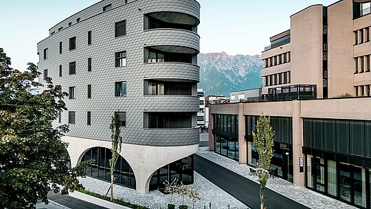 Vue de la Bernerhaus à Buchs, avec sa forme triangulaire remarquable et ses balcons aux courbes douces se détachant sur un fond de montagnes alpines. La façade continue en bardage PREFA gris souris P.10 enveloppe le bâtiment comme une peau d'aluminium aux allures d'écailles, créant un jeu d'ombre et de lumière saisissant. Au-dessus du socle en béton massif aux élégantes arcades, la façade légère et résistante aux intempéries en aluminium PREFA se distingue par sa précision, sa durabilité et son faible besoin d'entretien – un véritable atout architectural dans le paysage urbain.