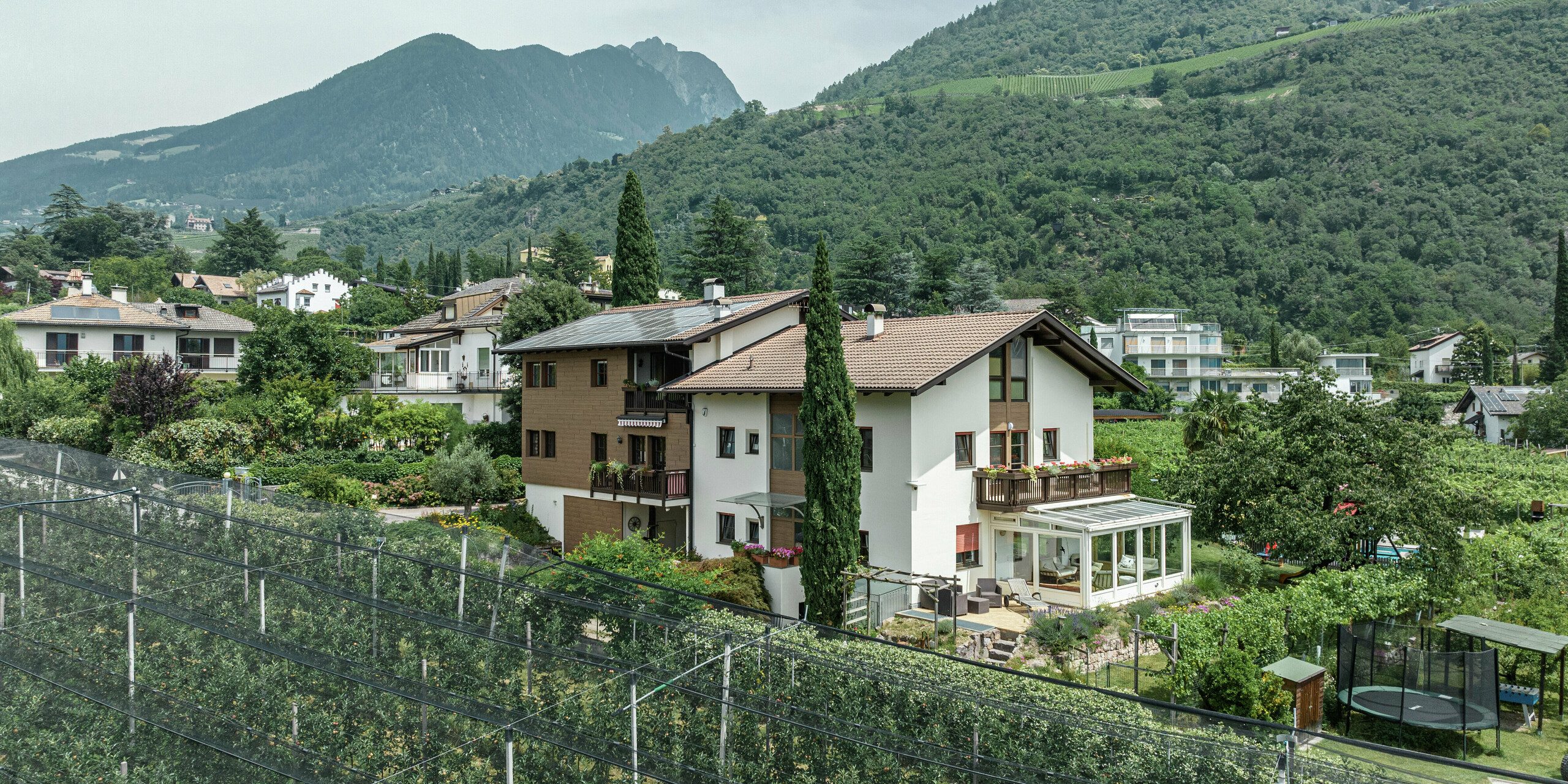 Vue aérienne des appartements de vacances Prechtlhof à Merano, dans le Tyrol du Sud, entourés de vergers et d'un paysage de montagne pittoresque. La façade a été modernisée avec des Sidings PREFA en noyer foncé, créant ainsi un aspect bois durable et sans entretien qui s'intègre harmonieusement dans l'environnement alpin.