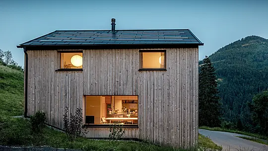 La maison en bois minimaliste Immi, à Ebnit, se détache sur le versant montagneux boisé dans la lumière du crépuscule. Sur le toit à deux pans aux lignes épurées, les panneaux solaires PREFA noirs se fondent presque imperceptiblement dans les R.16 en P.10 noir. La robuste couverture en aluminium, résistante aux intempéries, produit de l’énergie solaire, protège durablement le bâtiment et souligne l’architecture sobre et contemporaine de cette maison en bois alpine.