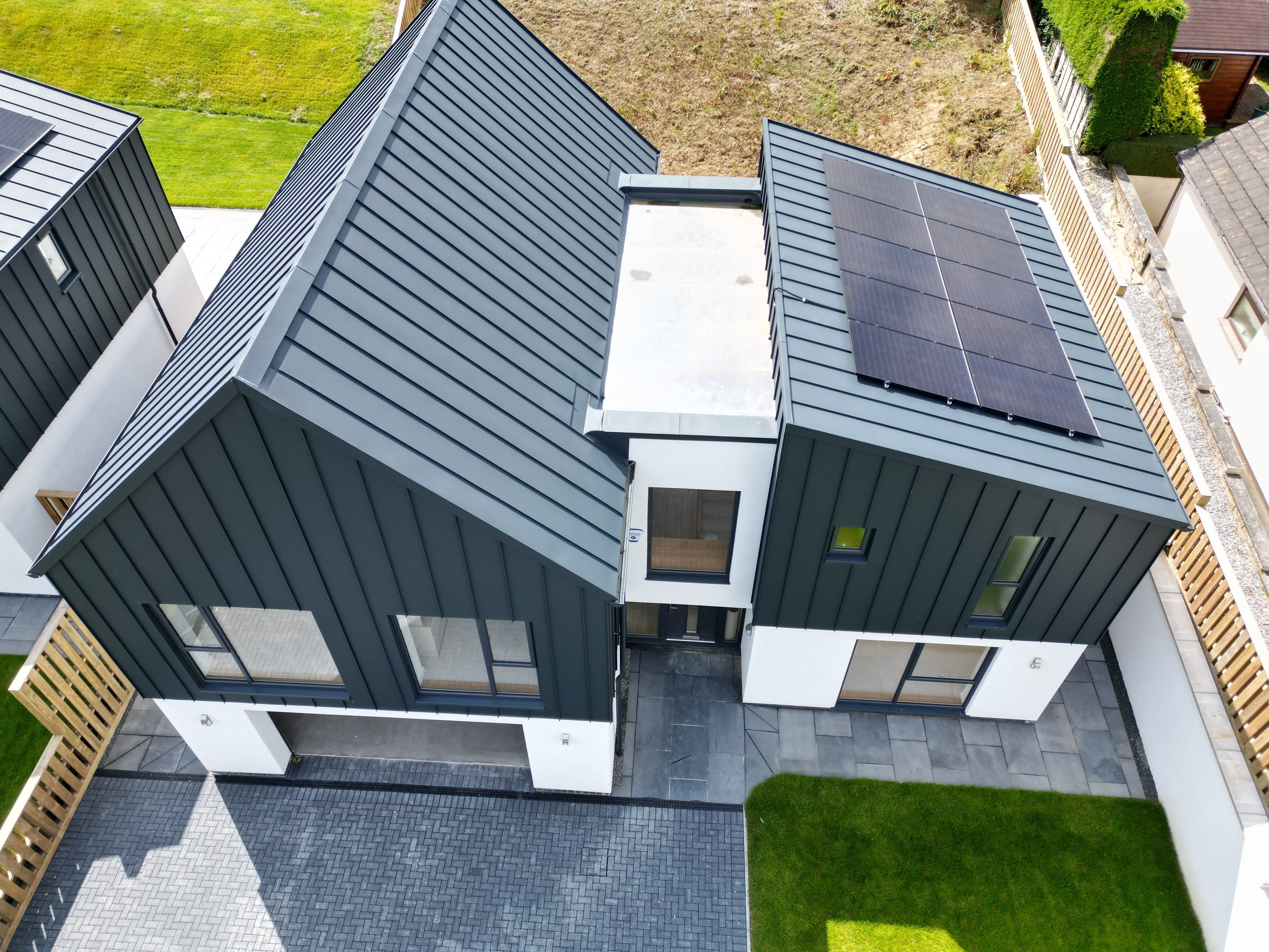 Vue aérienne d'une maison individuelle à Bridgerule, Devon, avec PREFALZ P.10 anthracite comme toiture à joint debout et façade. L'architecture moderne combine l'aluminium durable avec la longévité esthétique, complétée par un système solaire. Un toit à pignon domine l'entrée du garage et la partie droite du bâtiment est dotée d'un toit en appentis. Entre les toits, un pont-bâtiment relie les deux structures.